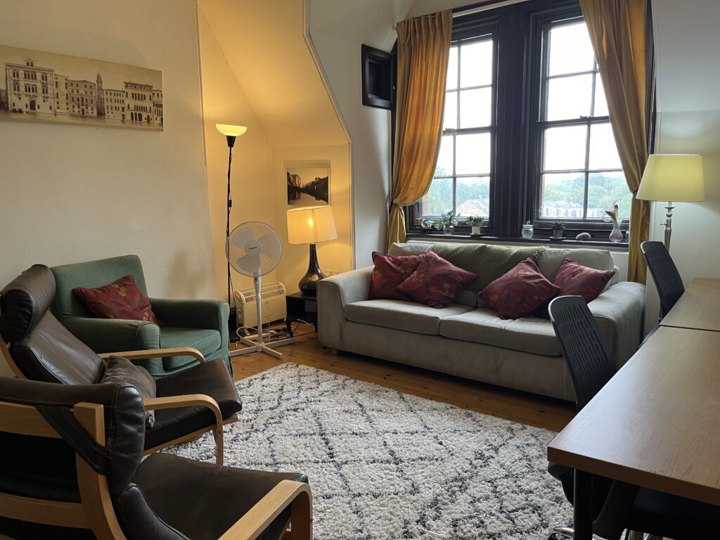 Image of interior of Library room space featuring lounge furniture, desk and window around a cosy rug and subdued lighting.