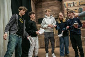 Five young actors stand in a row in rehearsal with scripts