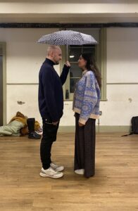 Male and female actor stand under un umbrella, smiling at each other