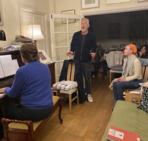 A male actor sings alongside a musician at a piano, with other actors looking on